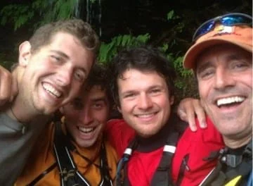 Four smiling people wearing outdoor gear, likely on a kayaking or hiking trip, posing for a close-up photo.
