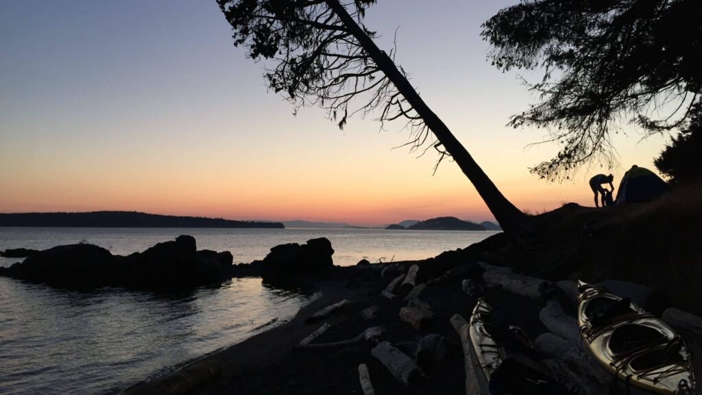 Scenic view of a shoreline at sunset with a silhouetted figure setting up a tent leaning trees kayaks on the ground and a calm body of water with distant islands under a colorful sky
