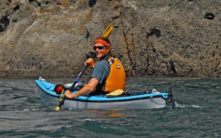 A person in a kayak wearing a life jacket and a headband smiling and paddling near a rocky shoreline