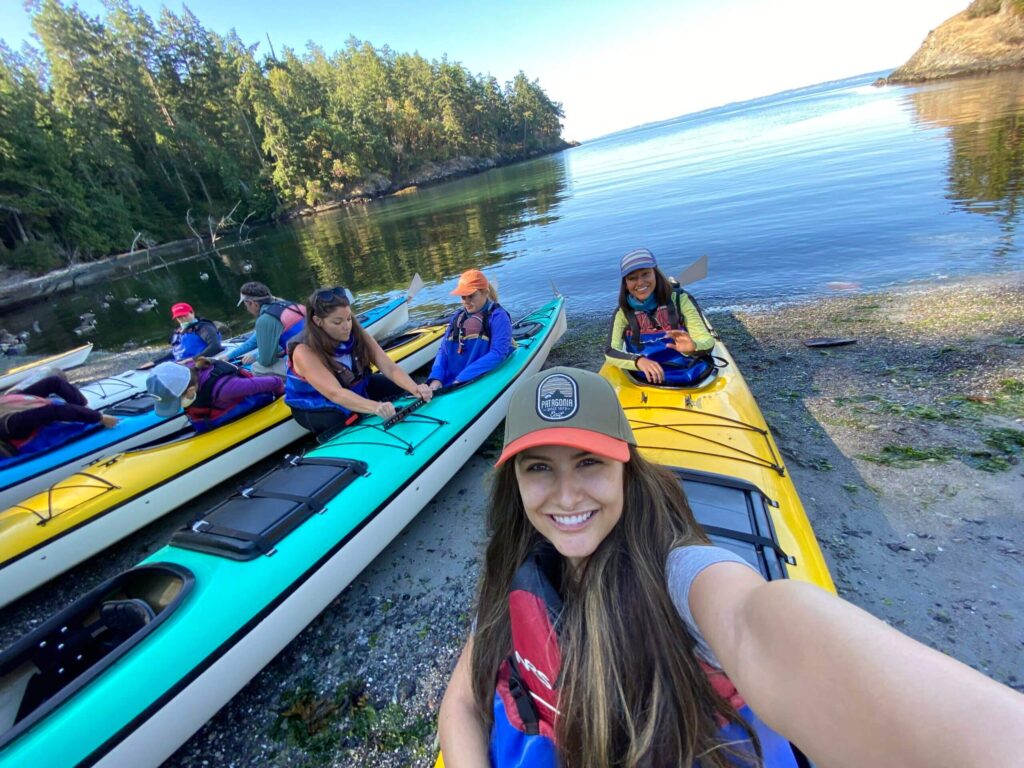 Group of people in colorful kayaks preparing for a kayaking trip with a woman in the foreground taking a selfie and smiling surrounded by a calm water inlet and dense forested area
