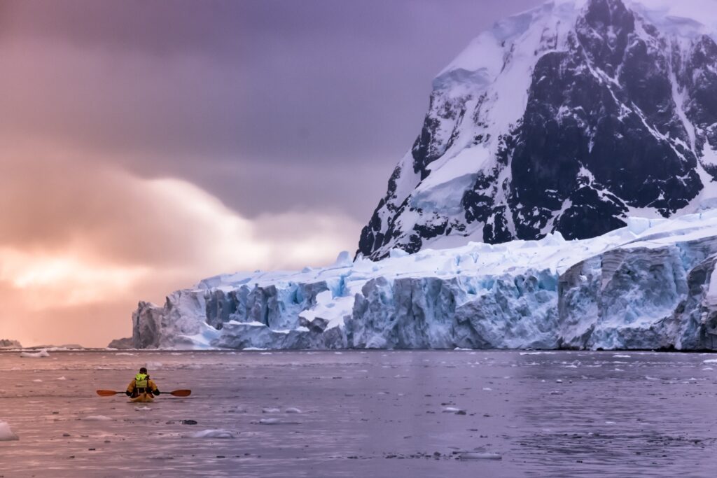 A person kayaking towards a massive snow covered glacier and mountain under a dramatic sky with hues of purple and orange