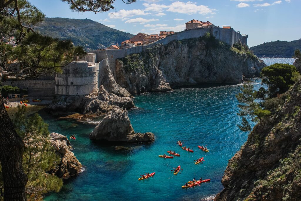 View of kayakers paddling in the clear turquoise waters near the rocky coastline of a historic town with stone fortifications and terracotta roofed buildings surrounded by lush greenery and mountains under a blue sky with scattered clouds