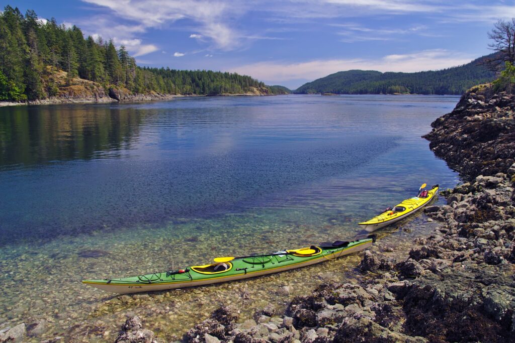 Two kayaks one green and one yellow rest on a rocky shore next to clear calm water surrounded by forested hills under a bright blue sky with wispy clouds