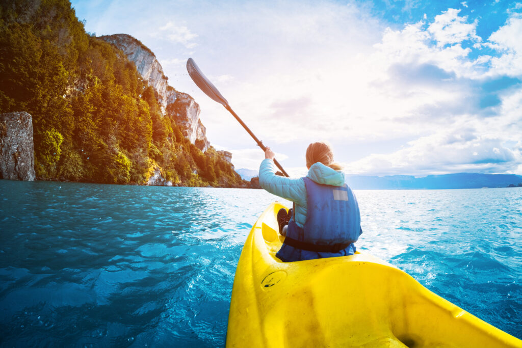 Person in a blue life jacket paddling a yellow kayak on a body of water with green rocky cliffs on the left and a bright sun shining through a partly cloudy sky