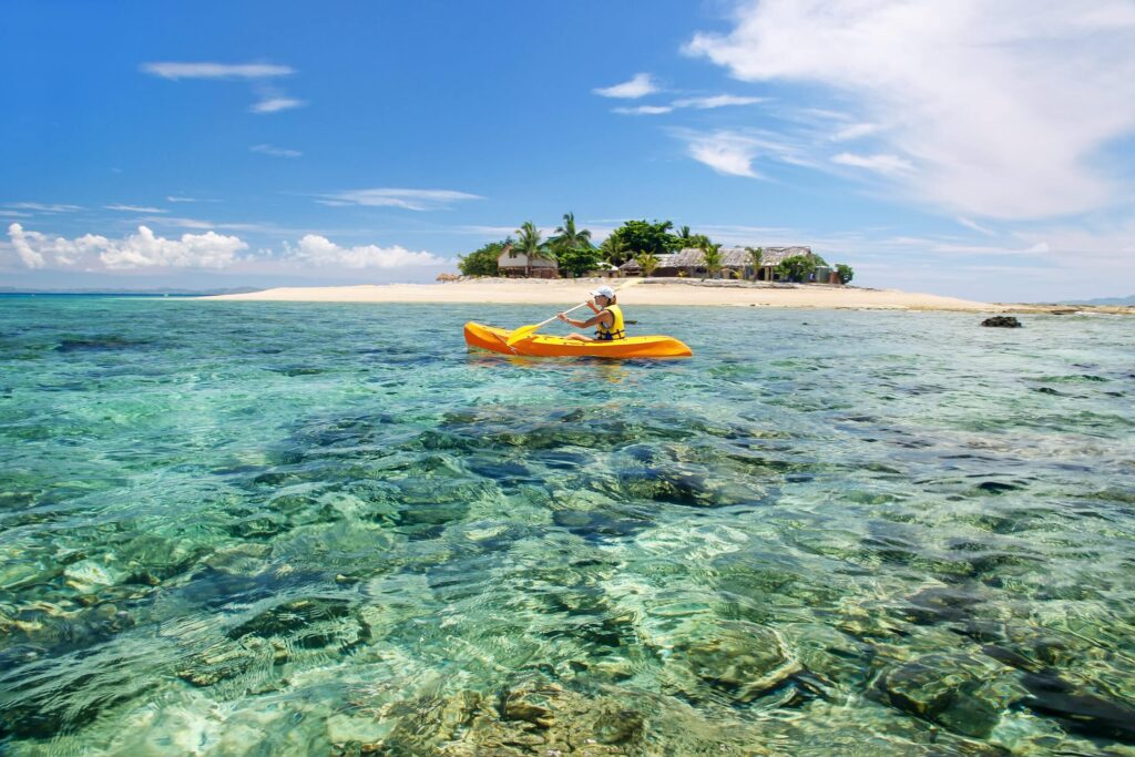 Person in a yellow kayak paddling on clear shallow water near a tropical island with white sandy beaches palm trees and huts under a bright blue sky with scattered clouds