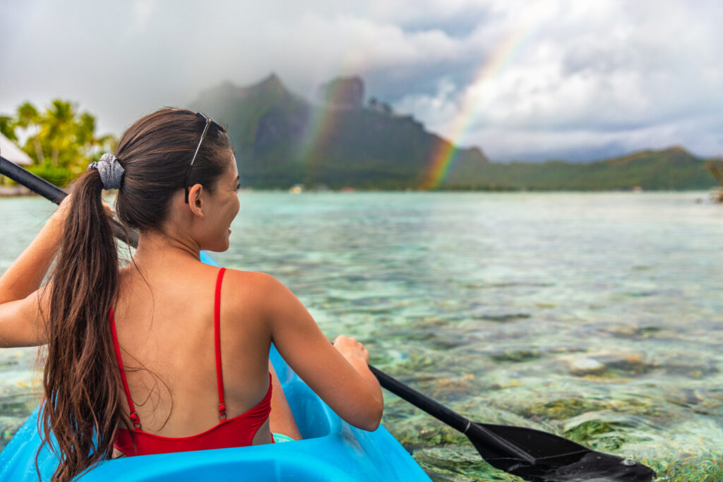 A woman in a red swimsuit paddling a blue kayak on clear shallow water with a rainbow in the sky and misty mountains in the background