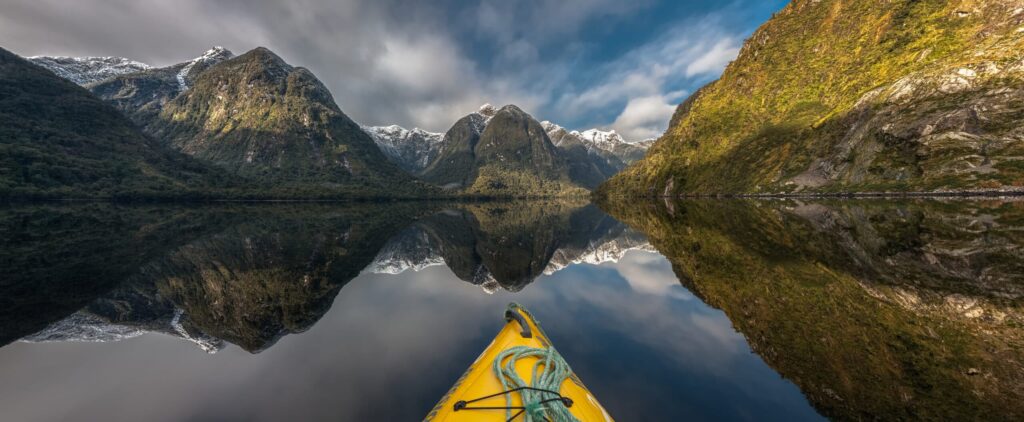 View from a yellow kayak on calm water surrounded by tall green mountains with snow capped peaks reflecting perfectly in the clear still water under a partly cloudy sky