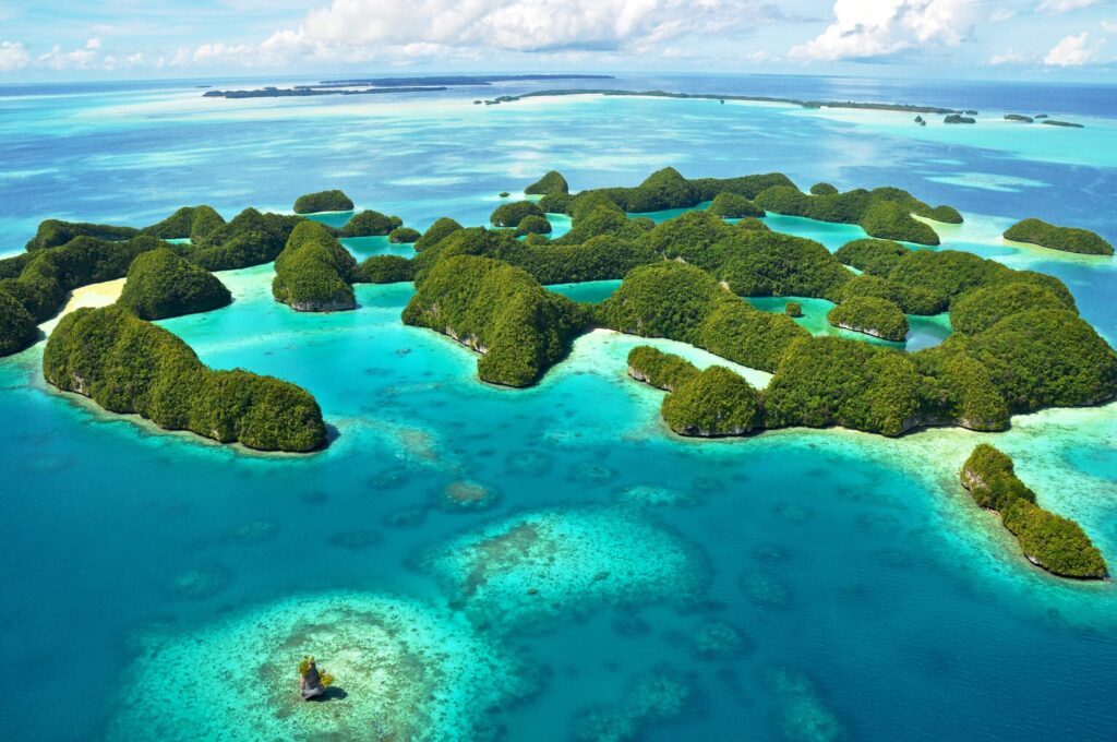 Aerial view of a group of lush green islands surrounded by turquoise waters and coral reefs with a larger expanse of blue ocean and distant islands under a partly cloudy sky
