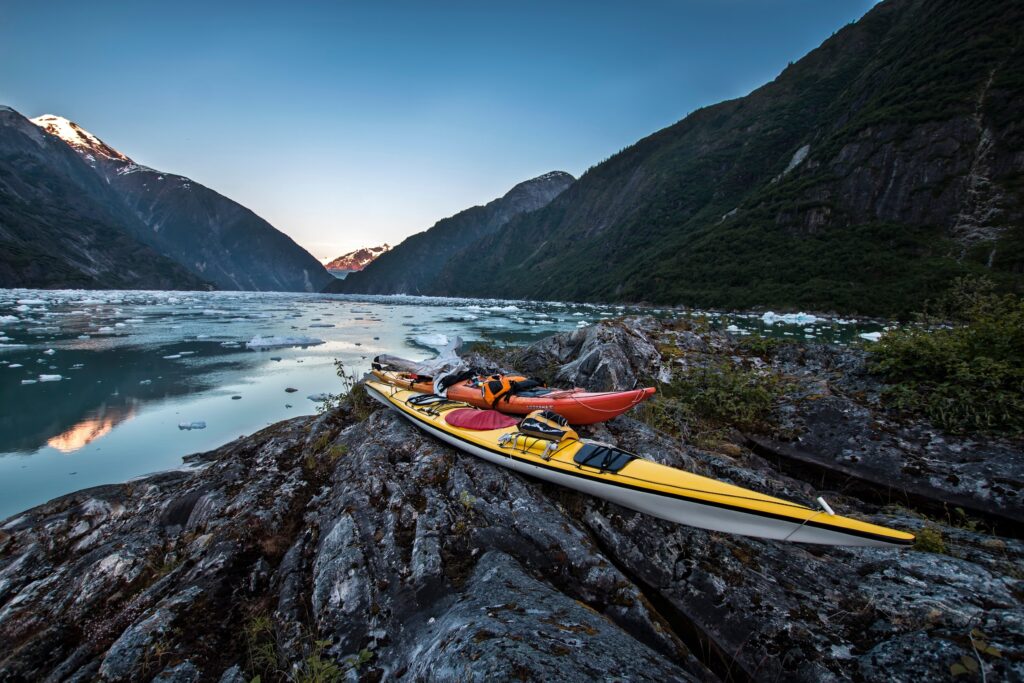 Two kayaks rest on rocky terrain overlooking a serene glacier lake surrounded by majestic snow capped mountains at dusk