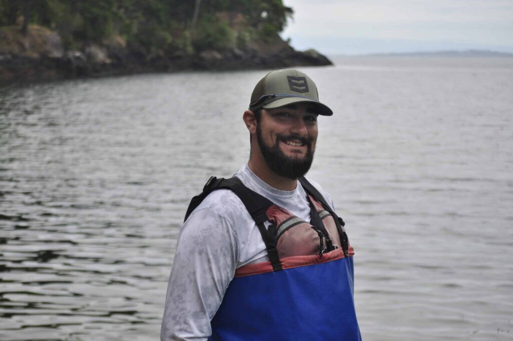Man smiling and standing near the water wearing a green cap a gray shirt and a red and blue life jacket with a rocky shoreline and trees in the background