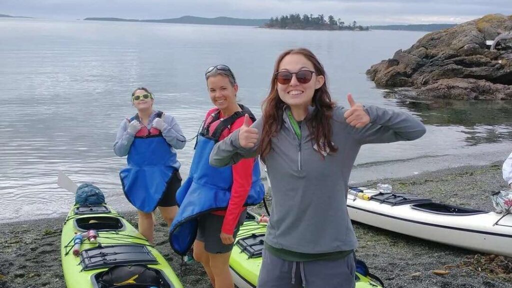 Three women standing on a beach next to kayaks all smiling and giving thumbs up Two of them are wearing blue spray skirts and life jackets and one is wearing sunglasses The calm sea and a rocky shoreline are visible in the background
