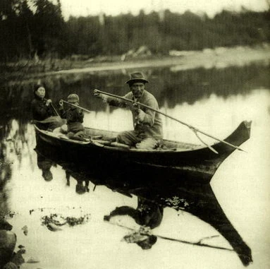 A historical black and white photo of a man a woman and a child fishing from a traditional wooden canoe on a calm river with a forested background