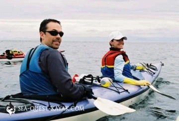 Two people sit in a tandem kayak on calm water They both wear life jackets and look back at the camera with the man in front wearing sunglasses and the woman behind him smiling