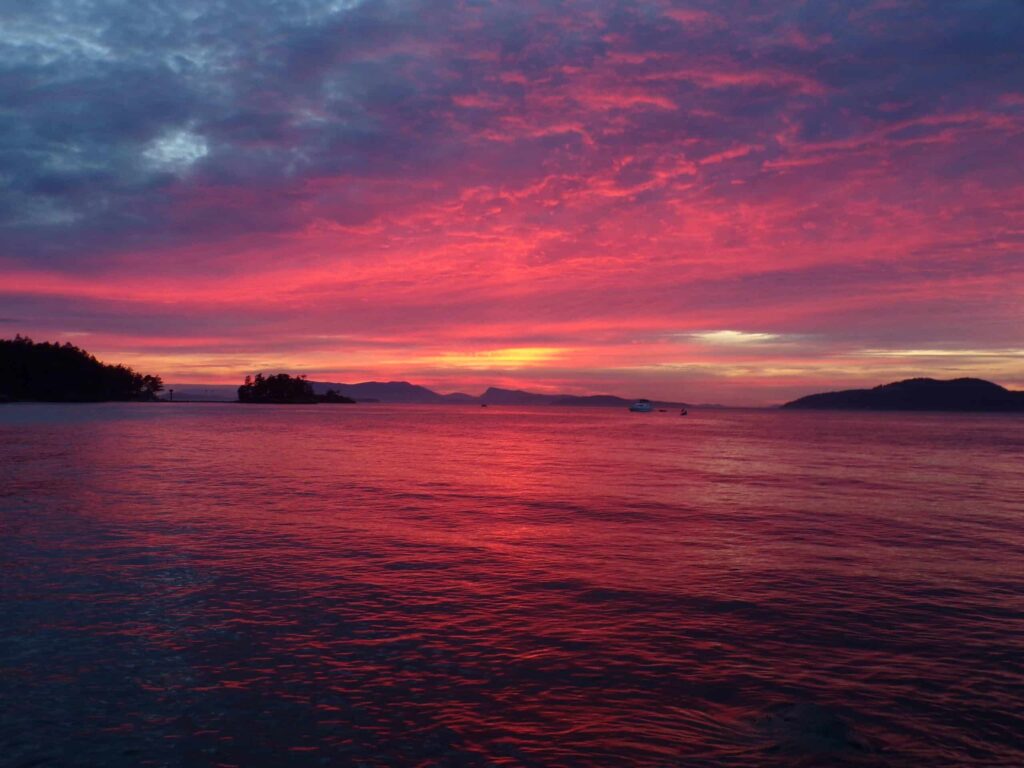 A stunning sunset over the water with vibrant pink purple and orange hues illuminating the sky and reflecting on the calm sea with silhouettes of islands and a boat in the distance