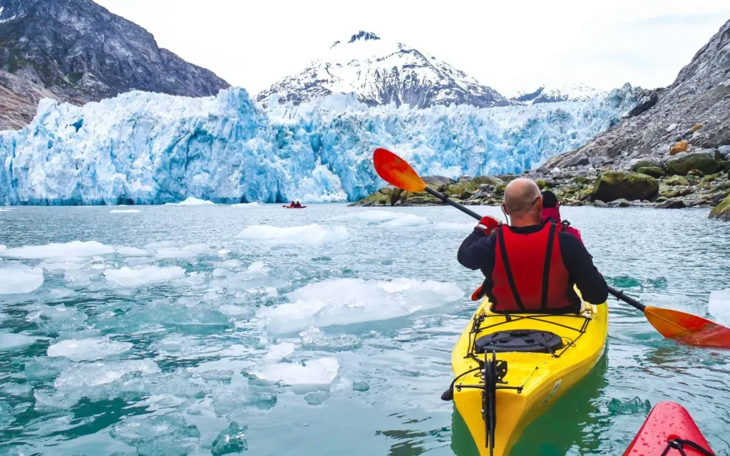 Person in a yellow kayak paddling towards a massive glacier surrounded by floating ice chunks and towering snow capped mountains