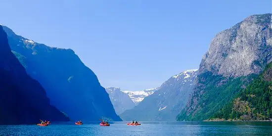 Group of kayakers paddling on a wide calm body of water surrounded by tall rugged mountains with patches of snow under a clear blue sky